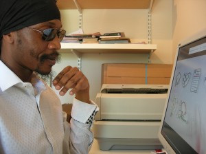 Picture of a black guy with a black headwrap, dark sunglasses and white shirt staring at the computer screen in an office.