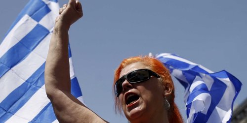 image of a woman with red hair, wearing dark sunglasses, waving a Greek white and blue flag and punching the air with a clenched fist.