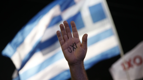 A hand is held up in front of a blue and white Greek flack in the background and a night sky. The inside of the palm is written in black in OXi which means NO!